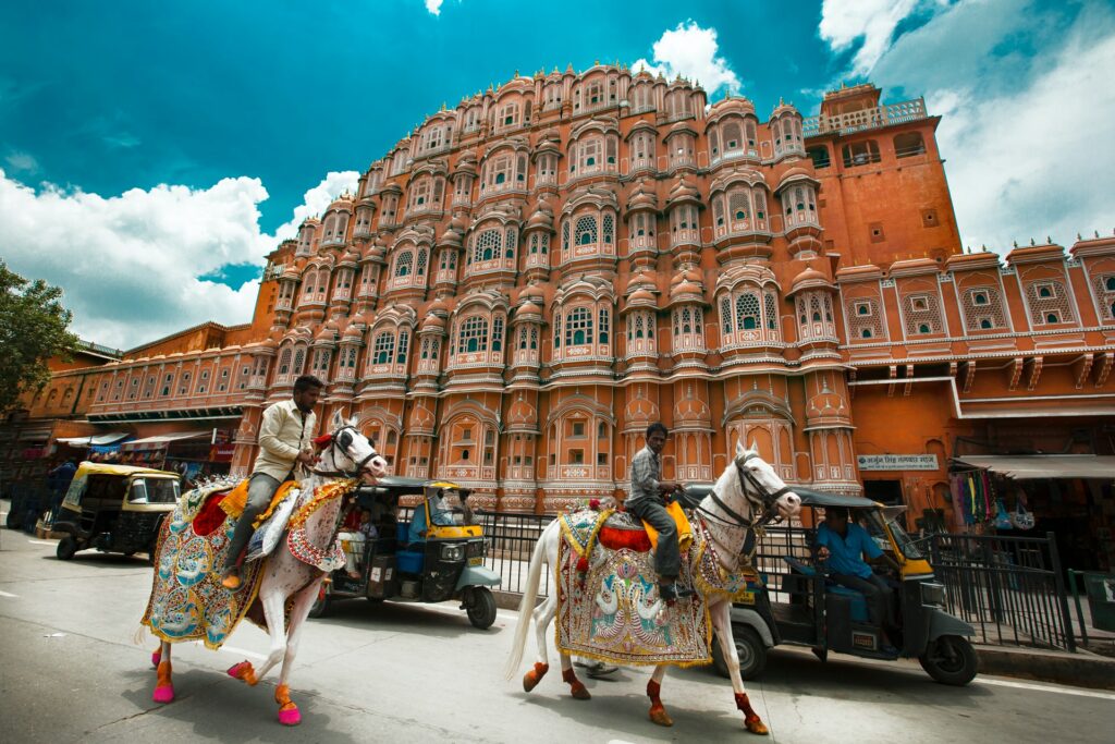 Hawa Mahal ou Palácio dos Ventos, em Jaipur, capital do estado de Rajastão na Índia, uma construção antiga com muitas janelas, em tom terroso, numa rua movimentada com carros e cavalos passando na frente do local, para representar o seguro viagem para Índia