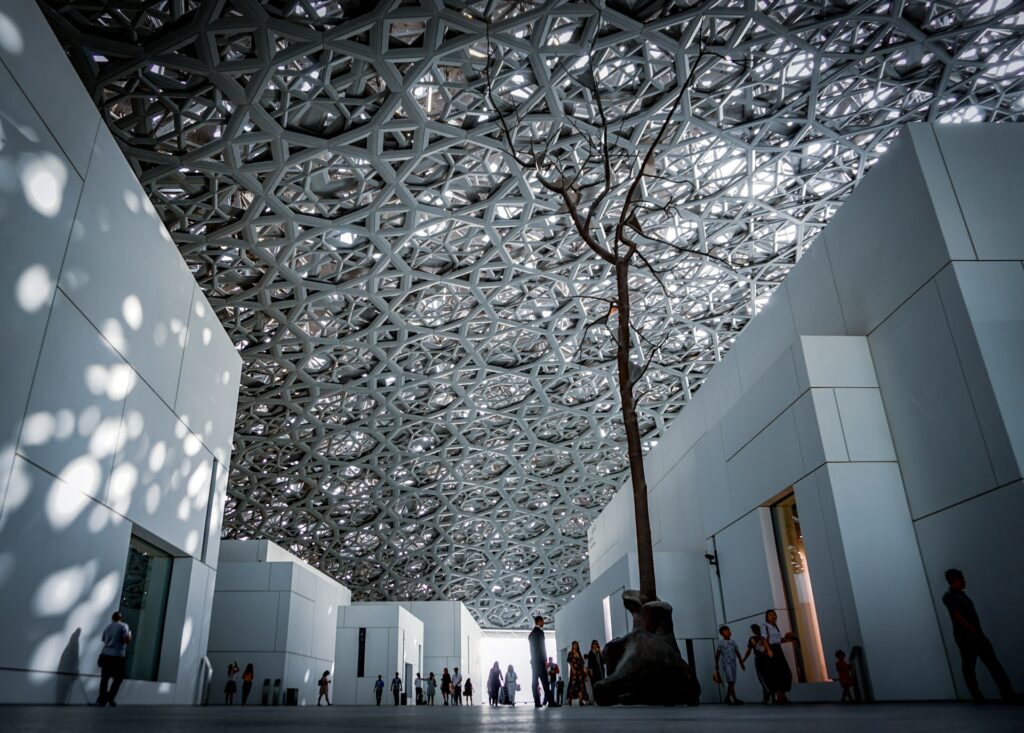 Museu Louvre Abu Dhabi com um teto que imita um emaranhado de galhos com a luz do sol passando entre eles, pessoas caminhando dentro do local que possuí muitas paredes brancas e é bem amplo, para representar o seguro viagem para os Emirados Árabes