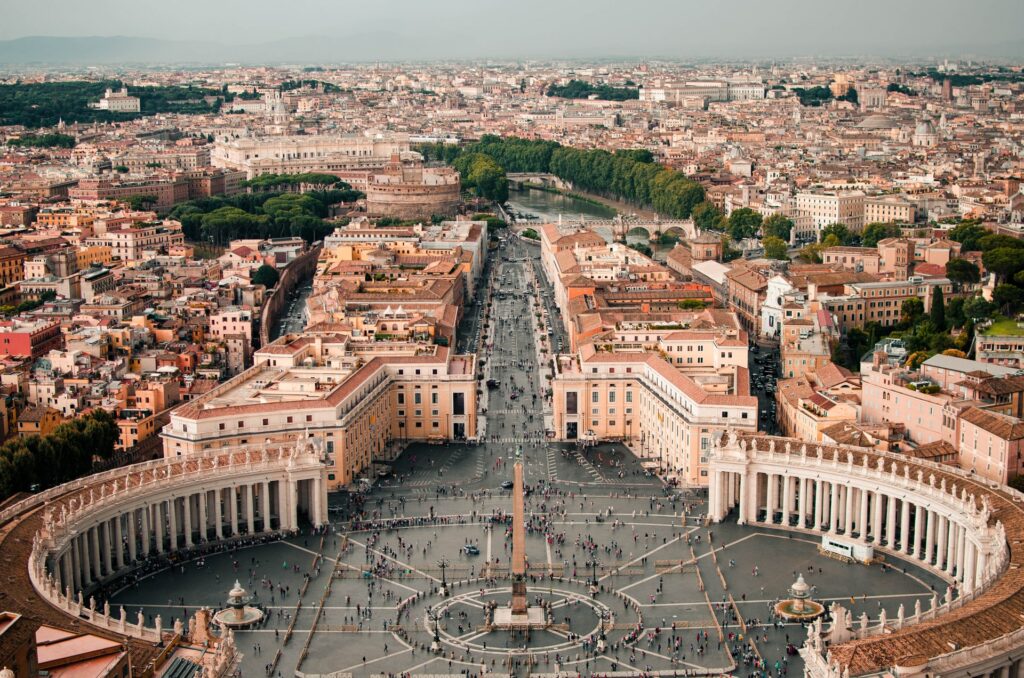 Praça redonda e ampla no Vaticano, local conhecido por ser o berço da igreja católica, com a cidade ao redor, um lago e árvores mais ao fundo para representar o seguro viagem para Roma