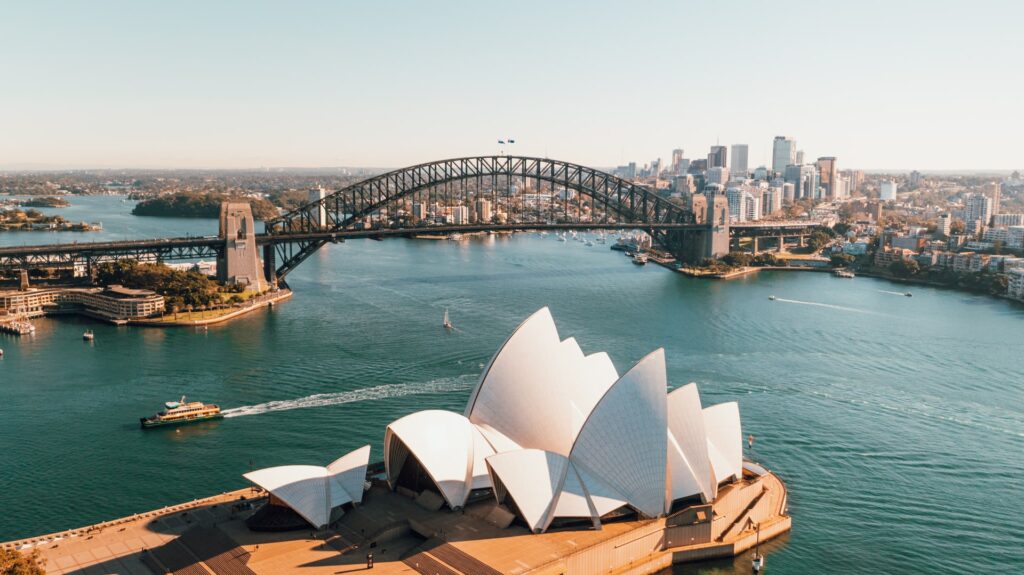Uma ponte passando por um enorme rio que interliga a cidade de Sydney na Austrália, barcos pequenos e grandes se locomovem, e também é possível ver o marco da cidade, a Opera de Sidney: um monumento de formas ondulares com pontas, para representar o seguro viagem para Austrália