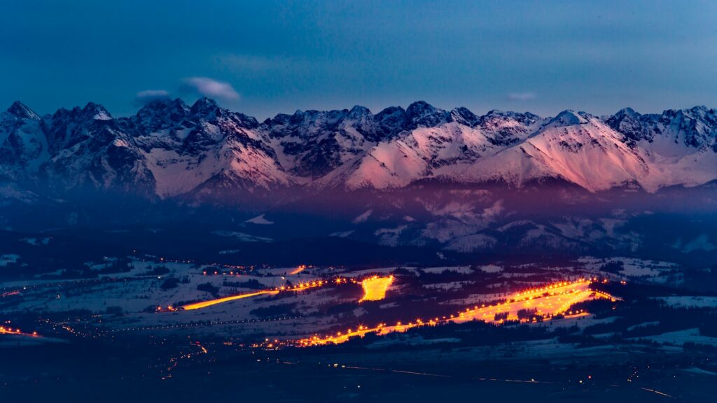 Montanhas nevadas com uma cidade com as luzes acesas ao pé desses picos, conhecido como  Montanhas Gorce, para representar o seguro viagem para Polônia