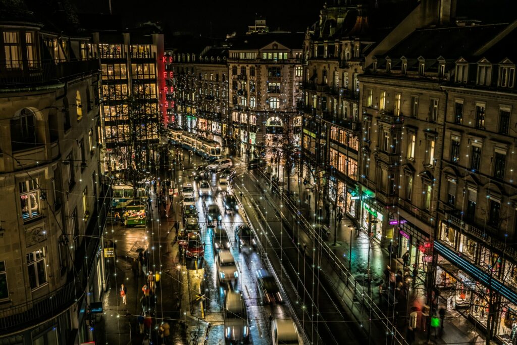 Zurique com as ruas iluminadas com luzes de natal presas nos prédios iluminando as ruas, carros passando, as fachadas das lojas também iluminadas, os prédios são antigos, para representar o seguro viagem para Zurique