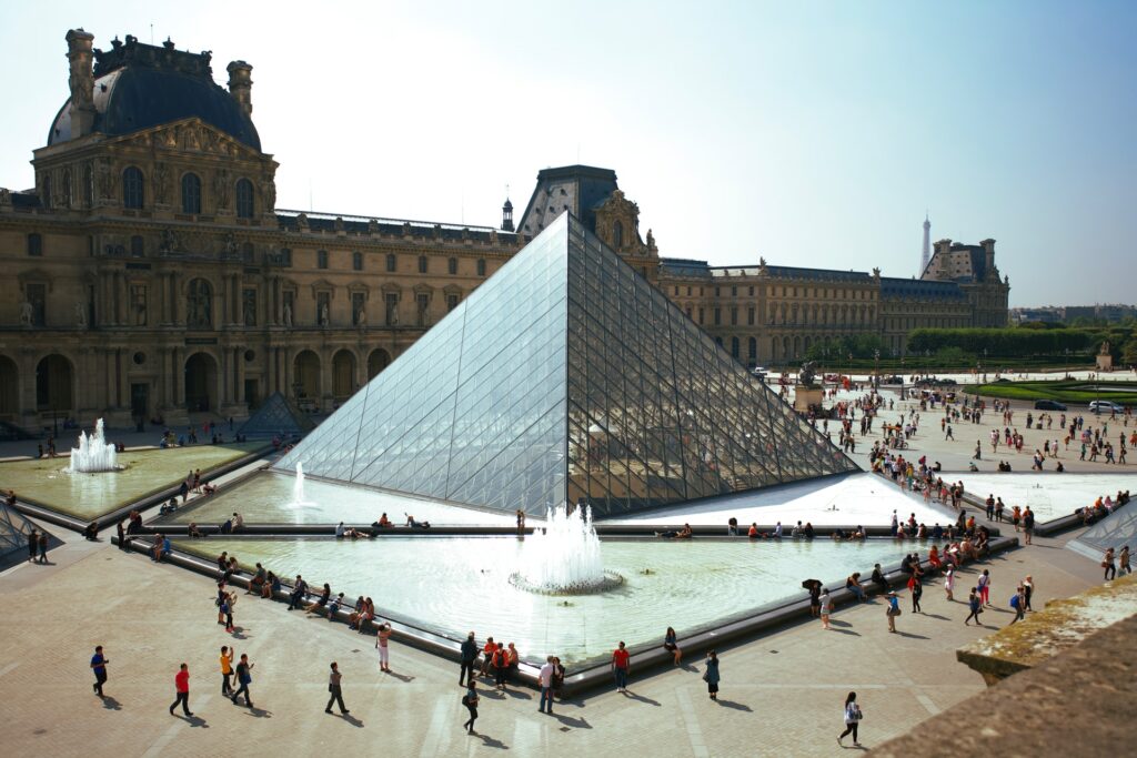 Praça com uma fonte e a pirâmide do Museu do Louvre logo atrás, pessoas circulando, prédio histórico ao fundo, para representar o seguro viagem para Paris