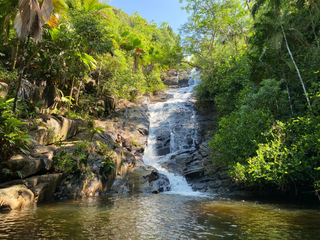 Uma cascata com águas límpidas em Port Glaud, cercado de rochas e vegetação, criando um lago ao fim da queda d'água, para representar o seguro viagem para Seychelles