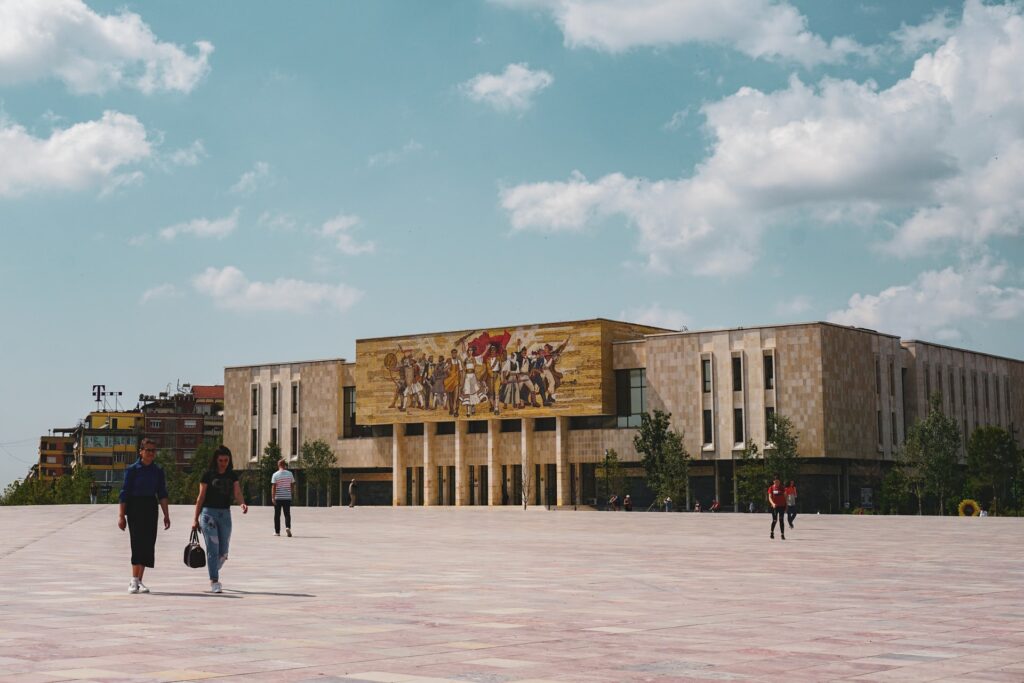 National History Museum em Tirana, na Albânia, fachada do extenso prédio, onde logo na entrada é possível ver uma pintura de cavaleiros e pessoas lutando, à frente do local, um enorme pátio com pessoas caminhando, para representar o seguro viagem para Albânia