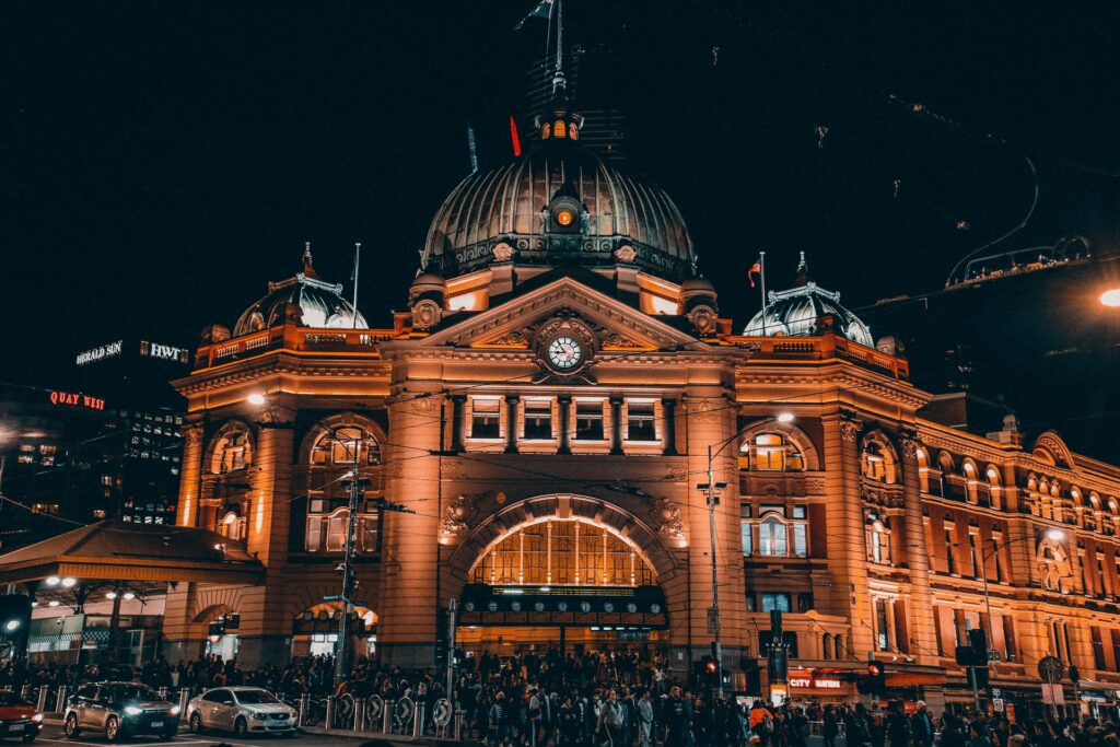 Flinders Street Railway Station em Melbourne na Austrália, uma construção antiga mantida, com um relógio ao centro e três cúpulas, com todas as luzes acesas de noite, para representar o seguro viagem para Austrália