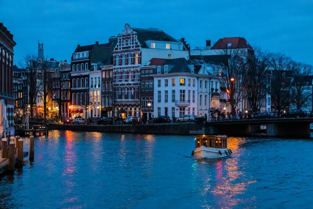 Amsterdam, a capital da Holanda, com a cidade beirando um lago com barcos passeando, uma ponte, casas em estilo antigo em tons de vermelho e branco, luzes acesas pelas ruas 