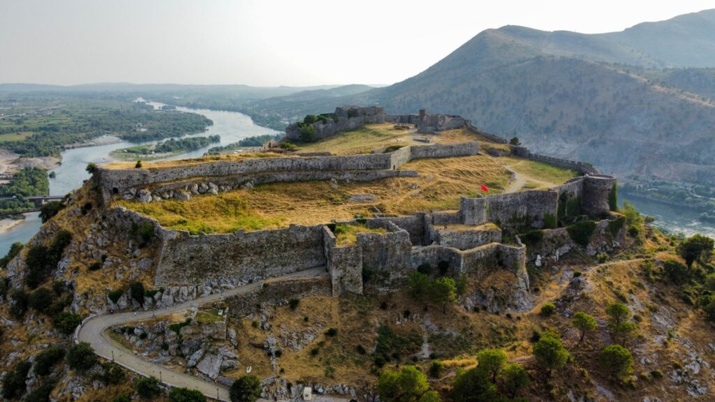 Castelo Rozafa na Albãnia, as ruínas de um castelo no topo  de uma montanha, cercado por vegetação, um rio e outras montanhas, para representar o seguro viagem para Albãnia