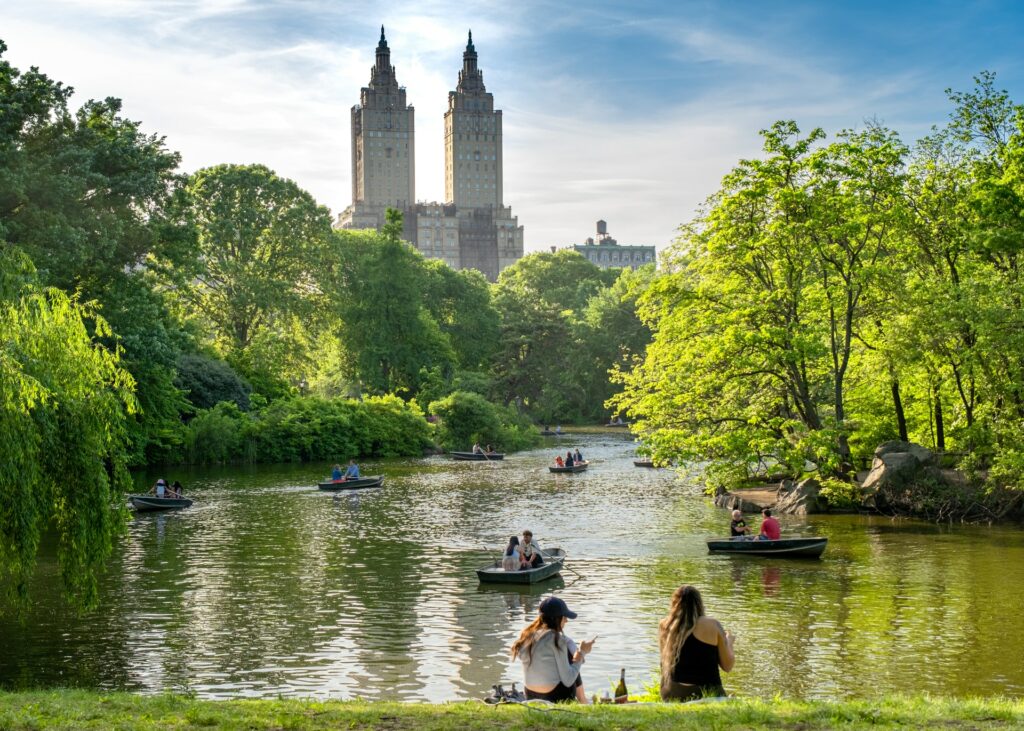 Um parque cercado de árvores, com um lago cheio de pequenos parquinhos e um gramado, ao fundo, prédios da cidade de Nova York para representar o seguro viagem para Nova York