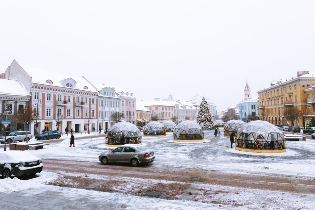 Cidade de Vilnius, com pequenas cúpulas de vidro presas numa praça, coberta por neve, prédios antigos e alguns carros na rua, para representar o seguro viagem para Lituânia