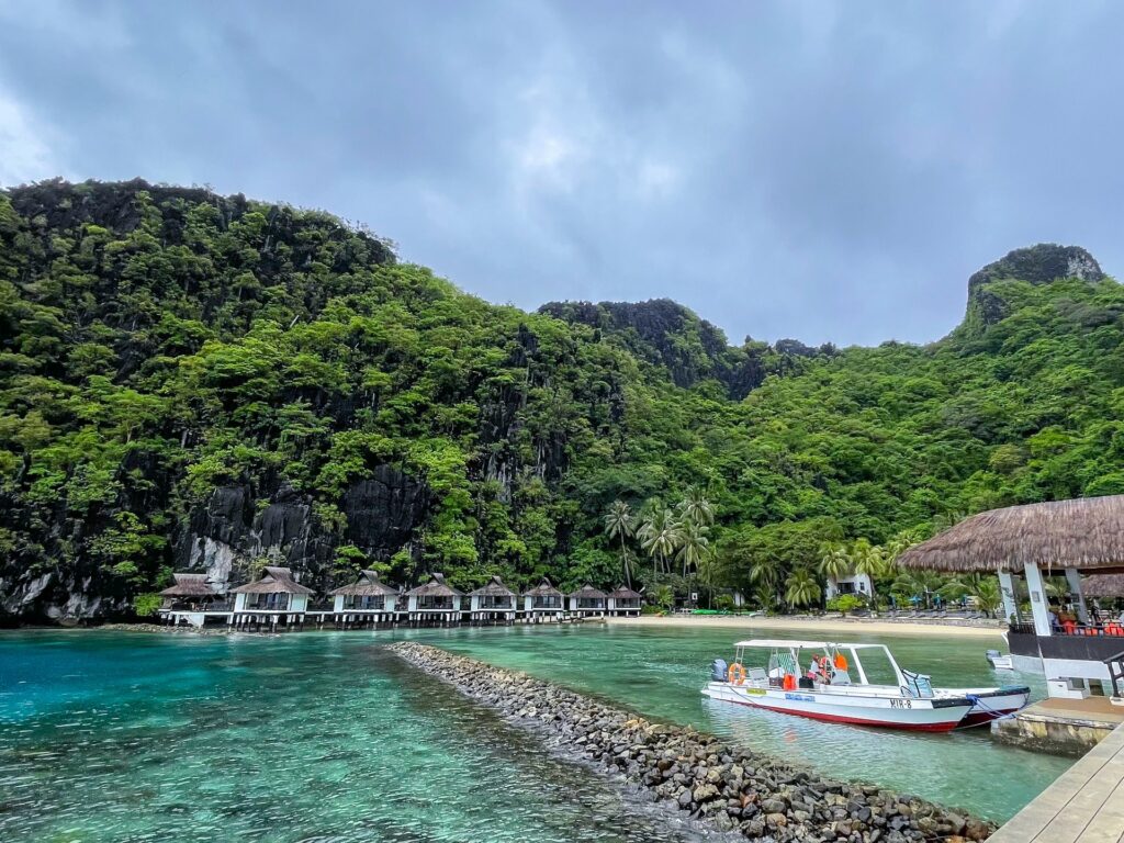 Ilha Miniloc com muitas montanhas cobertas de vegetação ao fundo, uma praia logo à frente, com águas transparentes, um quiosque, um barco e algumas casinhas que parecem estar flutuando no mar, para representar o seguro viagem para Filipinas