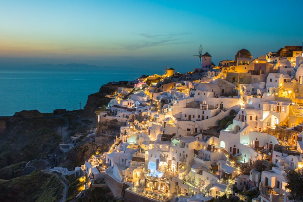 Diversas casas brancas construídas em uma rocha com as luzes acesas em Oia, uma cidade costeira da Grécia para representar o seguro viagem para Europa