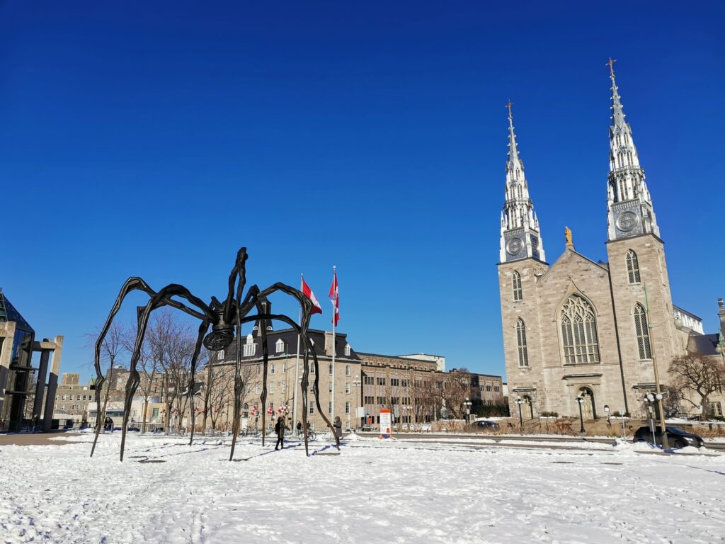 Prédios históricos, neve cobrindo as ruas, uma obra em metal que parece uma aranha em frente a Galeria Nacional do Canadá em Ottawa