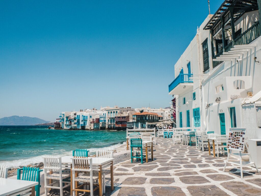Uma calçada com cadeiras brancas e azuis com o mar azul claro ao lado, casas nesse mesmo tom complementam a paisagem em Mýkonos na Grécia