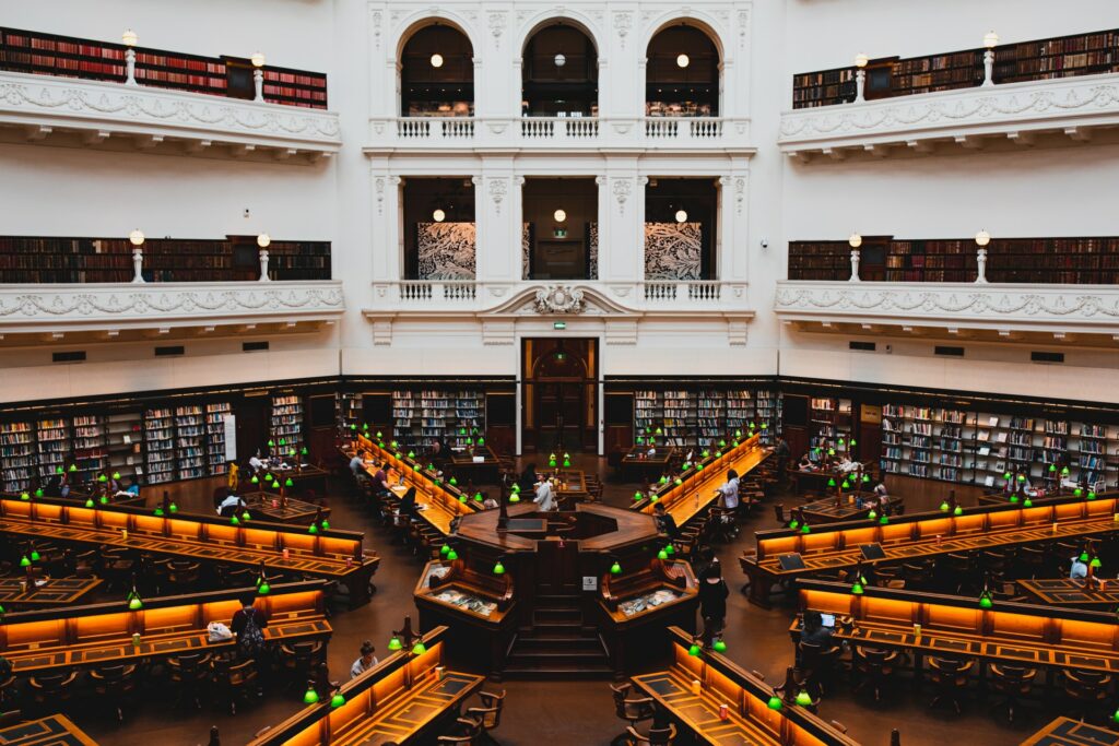 Parte interna da State Library de Victoria com uma arquitetura vitoriana, diversas estantes de livro e mesas iluminadas para sentar-se e ler um livro