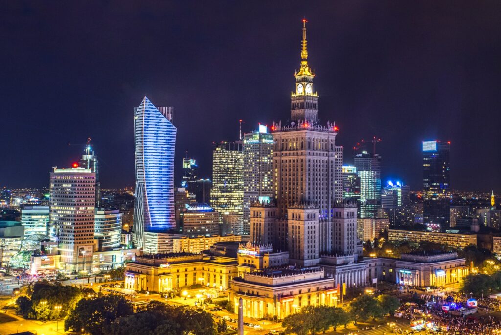 Varsóvia, capital da Polônia, inteira acesa de noite, com prédios iluminados, ruas cheias de pessoas, um enorme prédio com um relógio ao topo se destaca, muitas árvores pelas calçadas