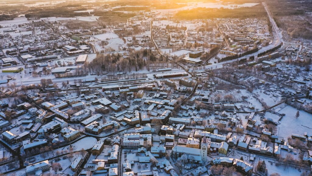 Cidade de Cesis na Letônia, coberta por neve, muitas casas com arquitetura antiga, uma torre de uma catedral, um pouco de vegetação e uma linha de trem cruzando a cidade, para representar o seguro viagem para Letônia