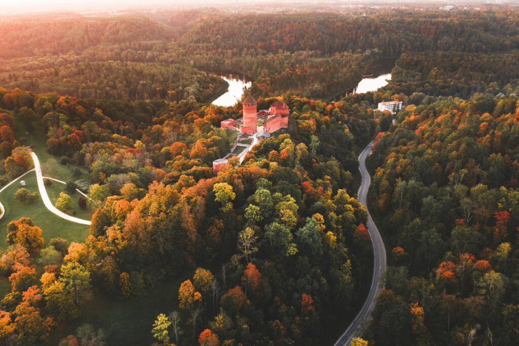 Cidade de Sigulda na Letônia com uma enorme floresta com árvores em tons de verde claro, escuro e laranja, com alguns lagos entre a vegetação, e um castelo escondido por entre a floresta, para representar o seguro viagem para Letônia