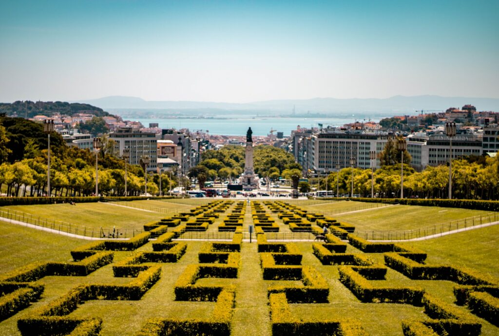 Um parque amplo, com jardins cheios de recortes, um monumento e o mar ao fundo, depois dos prédios da cidade, em Lisboa para representar o seguro viagem para Lisboa