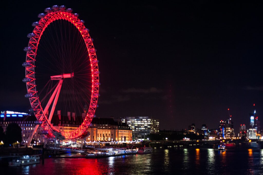 A roda gigante London Eye acessa em tons de vermelho de noite com a cidade acesa ao fundo e as cores refletindo na água 
