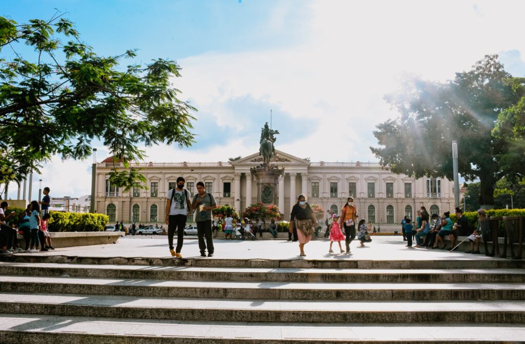 Praça Gerardo Barrios com o Palácio Nacional de El Salvador ao fundo com pessoas caminhando, conversando, alguns bancos próximos de arbustos, para representar o seguro viagem para El Salvador