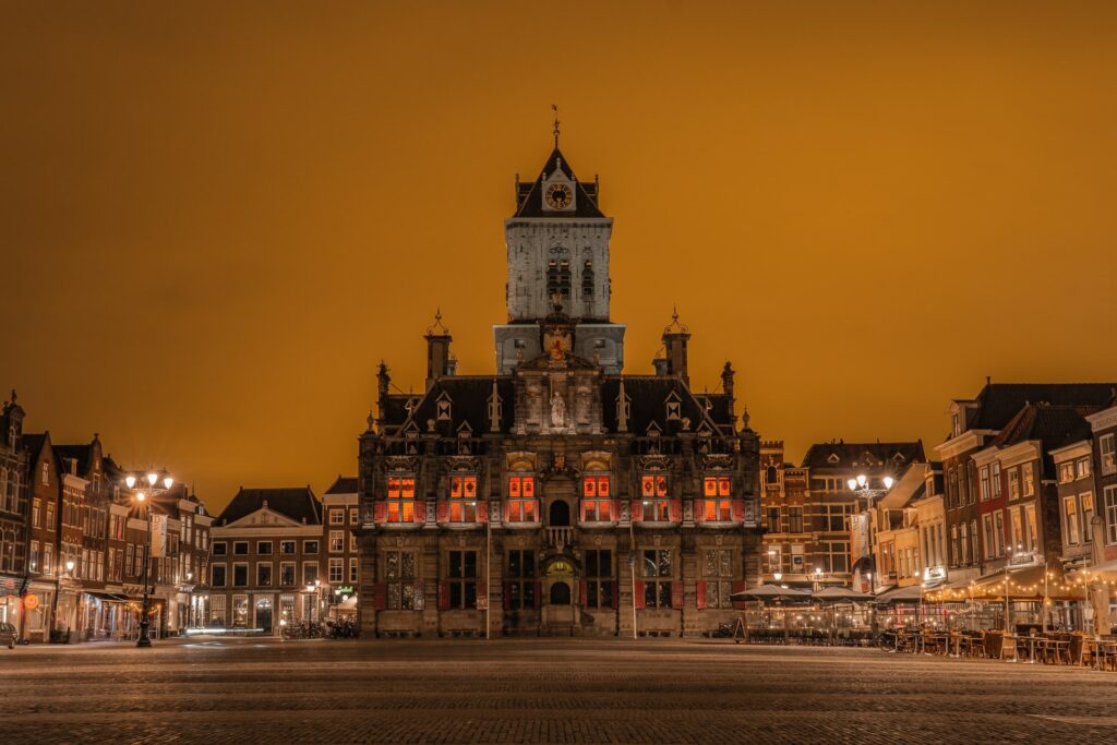 Prédio histórico com arquitetura colonial e barroca que abriga a prefeitura de Delft na Holanda, construção com três andares, mais uma torre que abriga um relógio, tudo em tons de marrom e laranja, para representar o seguro viagem para Holanda