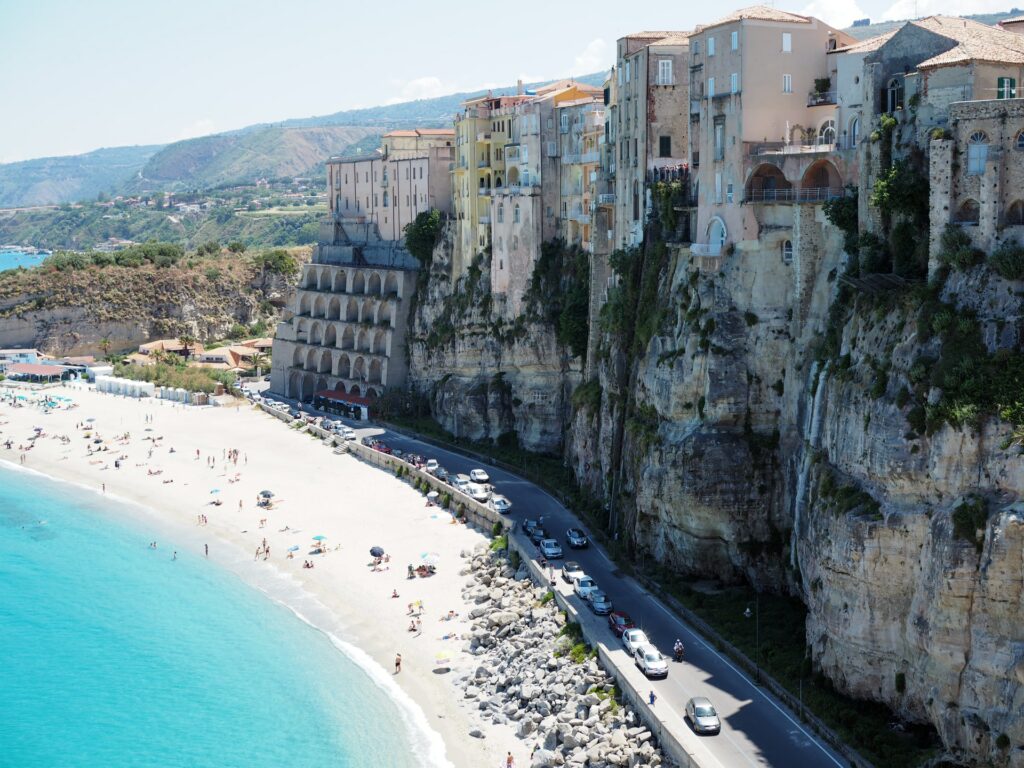 Rochas altas fazendo sombra numa pequena rua que está paralela com a areia da praia, águas transparentes e areia branca em Tropea, pequena cidade na costa lesta da Itália