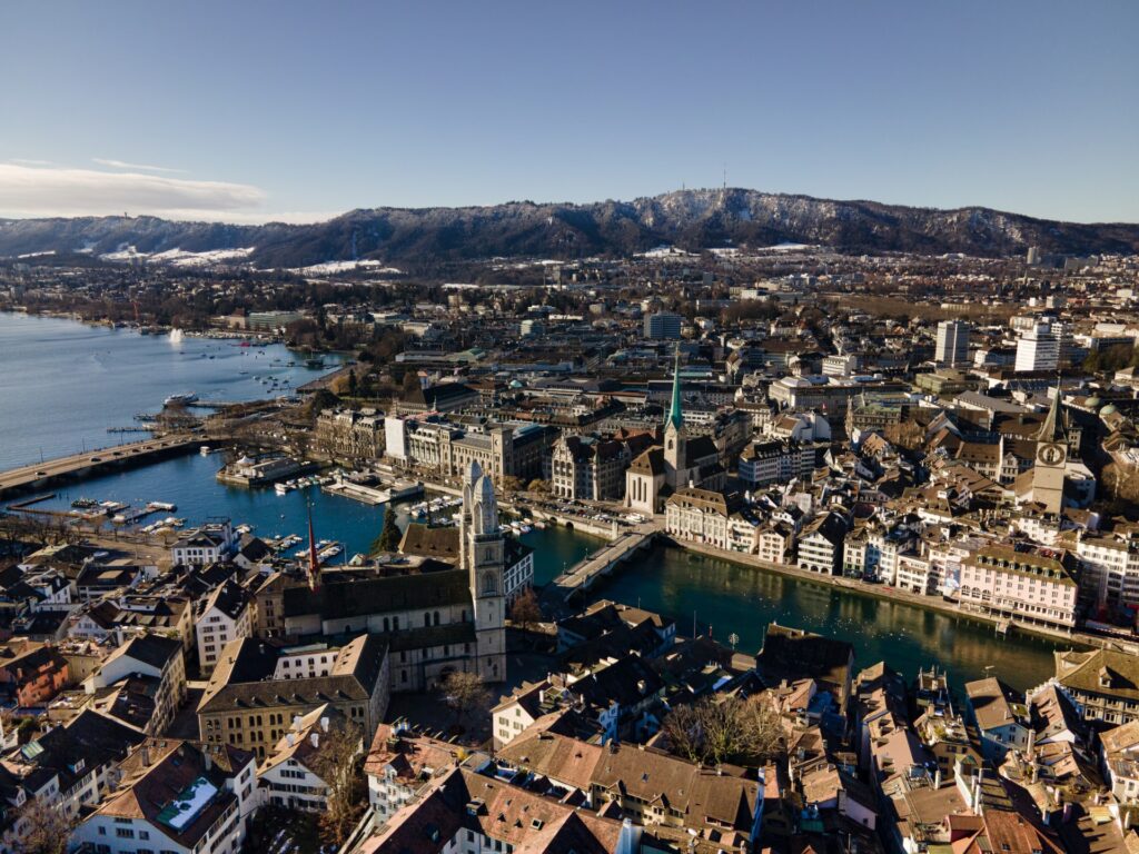 Distrito de Altstadt em Zurique, uma área com um rio, uma ponte que interliga os territórios, torres de catedrais antigas se destacam, muitas casas em arquitetura medieval e, ao fundo, montanhas nevadas, para representar o seguro viagem para Zurique