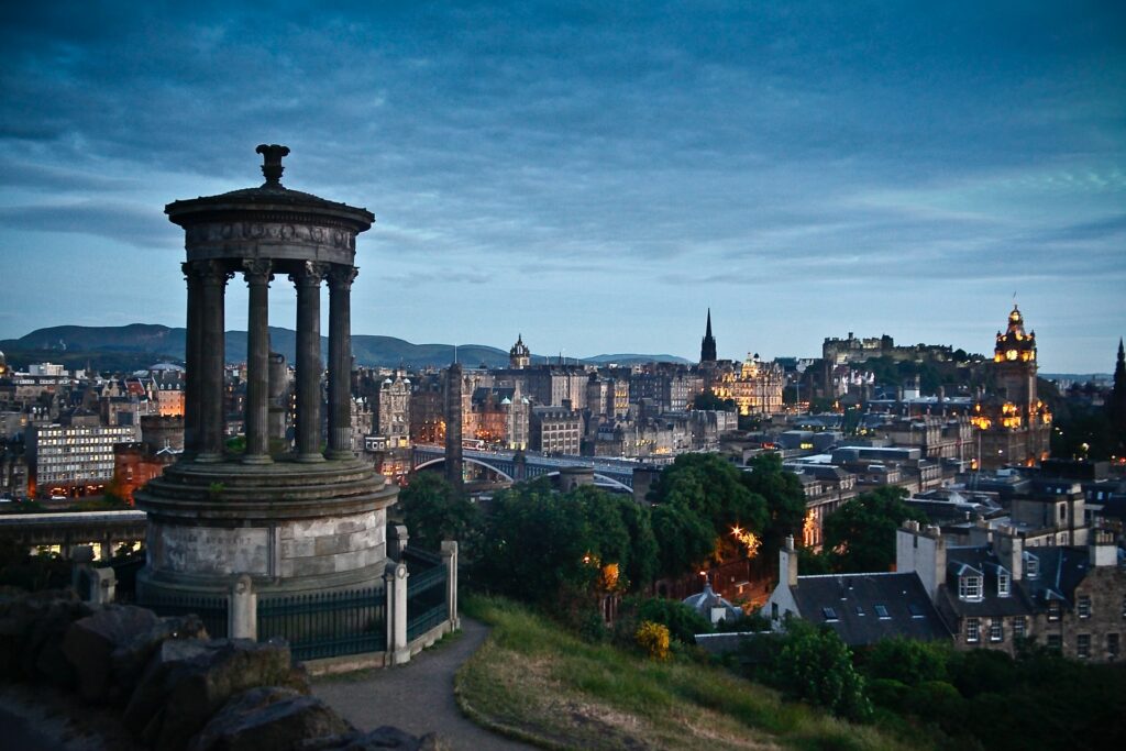 Centro de Edimburgo na Escócia de noite, diversas torres históricas estão as luzes acesas, casas em estilo antigo, árvores e monumentos se misturam e complementam a paisagem, para representar o seguro viagem para o Reino Unido