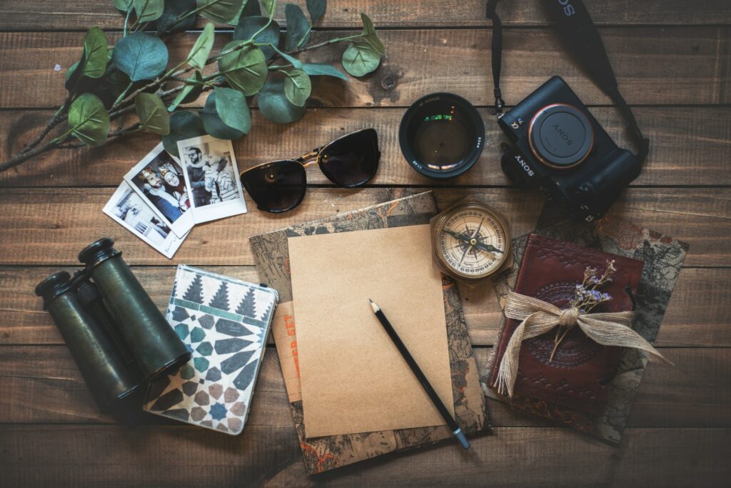 Uma mesa de madeira com um caderno, um lápis, uma bússola, um binóculo, um óculos de sol e algumas fotografias, para representar o seguro viagem para Polinésia Francesa