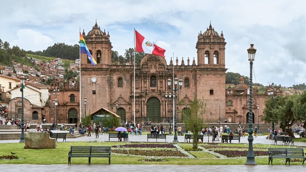 Uma praça florida com a bandeira do Peru hasteada e, ao fundo, um prédio histórico em tons terrosos com duas torres para representar seguro viagem em Lima