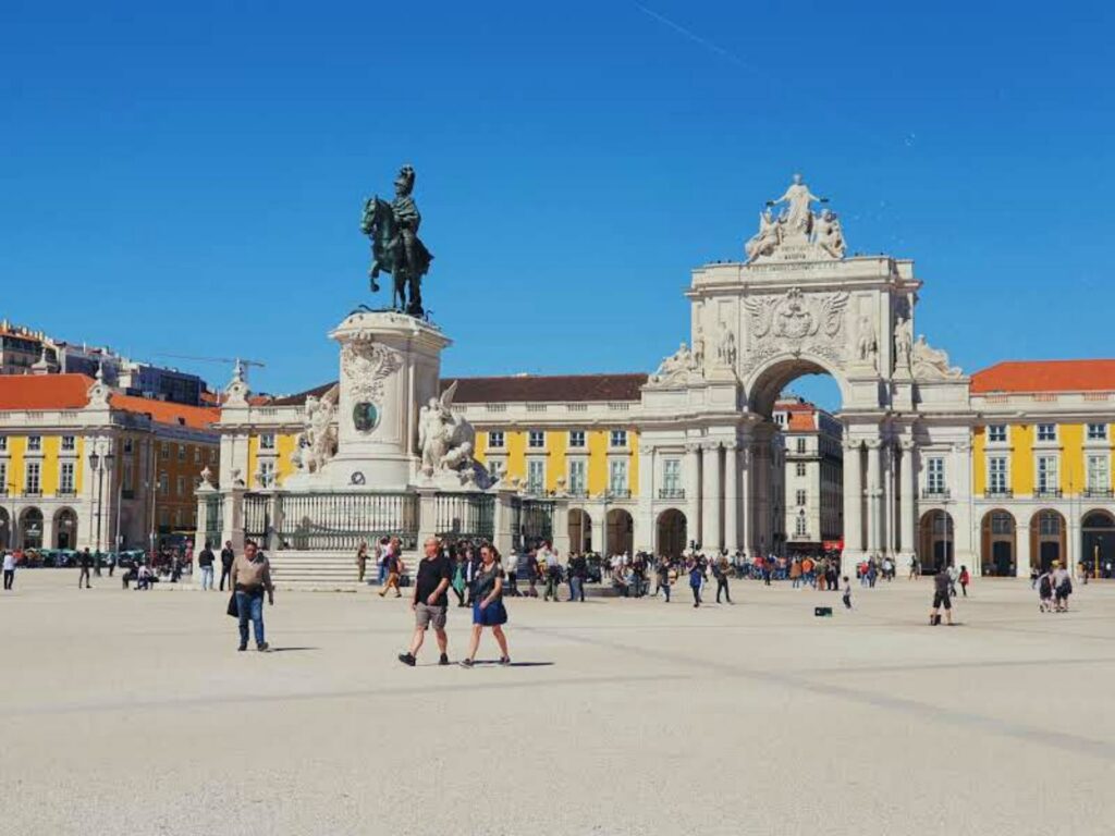Praça do Comércio, lugar amplo no centro da cidade de Lisboa com construções antigas em formato de arco, prédios históricos amarelos, um monumento de um homem sentado num cavalo, pessoas andando, para representar o seguro viagem para Lisboa