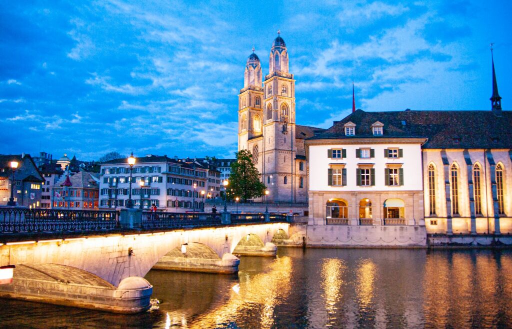Igreja Grossmünster em Zurique, uma ponte iluminada que leva até a igreja e outros prédios com arquitetura colonial e medieval, postes com luzes amarelas iluminam as ruas, para representar o seguro viagem para Zurique