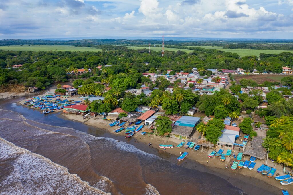 A costa de uma praia em Masachapa no Nicarágua, com muita vegetação, árvores e algumas casas perto de uma enorme área verde, à frente, o mar e alguns barcos parados, para representar o seguro viagem Nicarágua