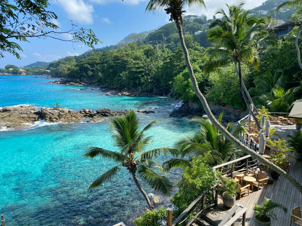 Muitos coqueiros, um mar límpido ao ponto de podermos enxergar as paredes de corais ao fundo, algumas rochas, muita vegetação e uma pousada virada para o mar em Praslin em Seychelles