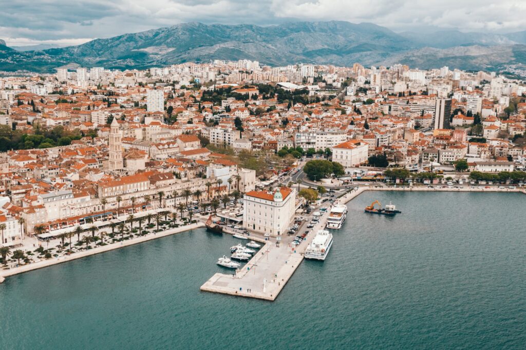 Uma cidade com montanhas ao fundo, prédios antigos, um porto que dá para o mar, com muitos barcos parados, na cidade de Split na Croácia
