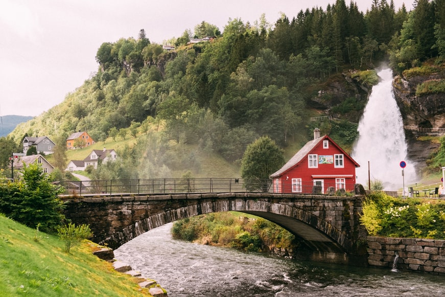 Casa de madeira em meio a uma ponte e uma cachoeira em Steinsdalsfossen, Norheimsund, Noruega - Representa seguro viagem Noruega.