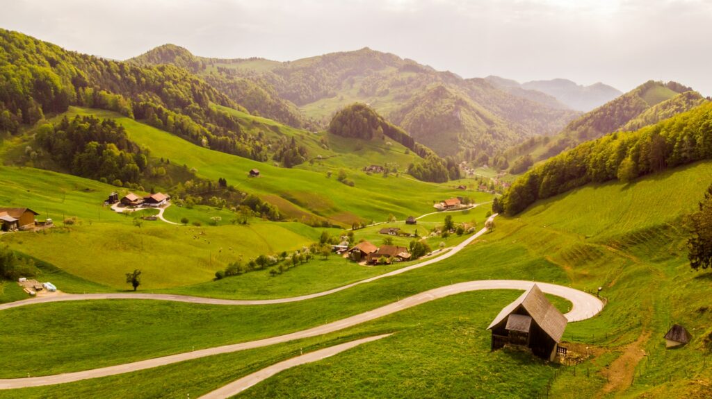 Um vasto campo com montanhas ao fundo, muitas árvores, algumas casas de madeira entre as colinas em Mümliswil-Ramiswil, uma comuna da Suíça