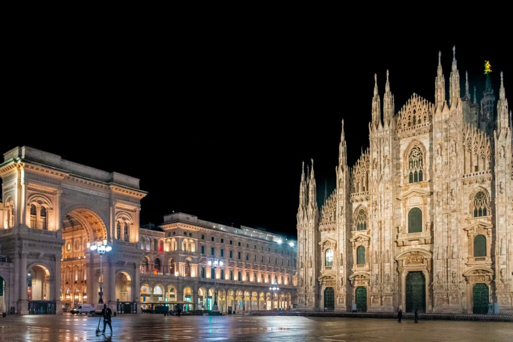 Piazza Del Duomo em Milão com construções antigas e com arquitetura romana, incluindo uma igreja e um monumento em formato de arco para representar o seguro viagem para Itália