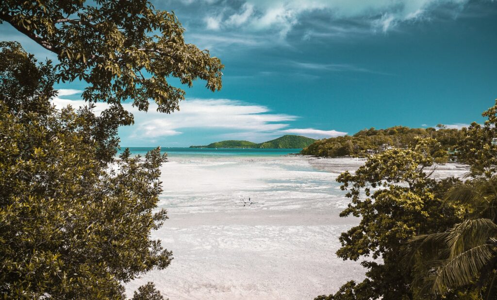 Praia com extensa faixa de areia branca, mar transparente, árvores e algumas montanhas ao fundo no Taiti