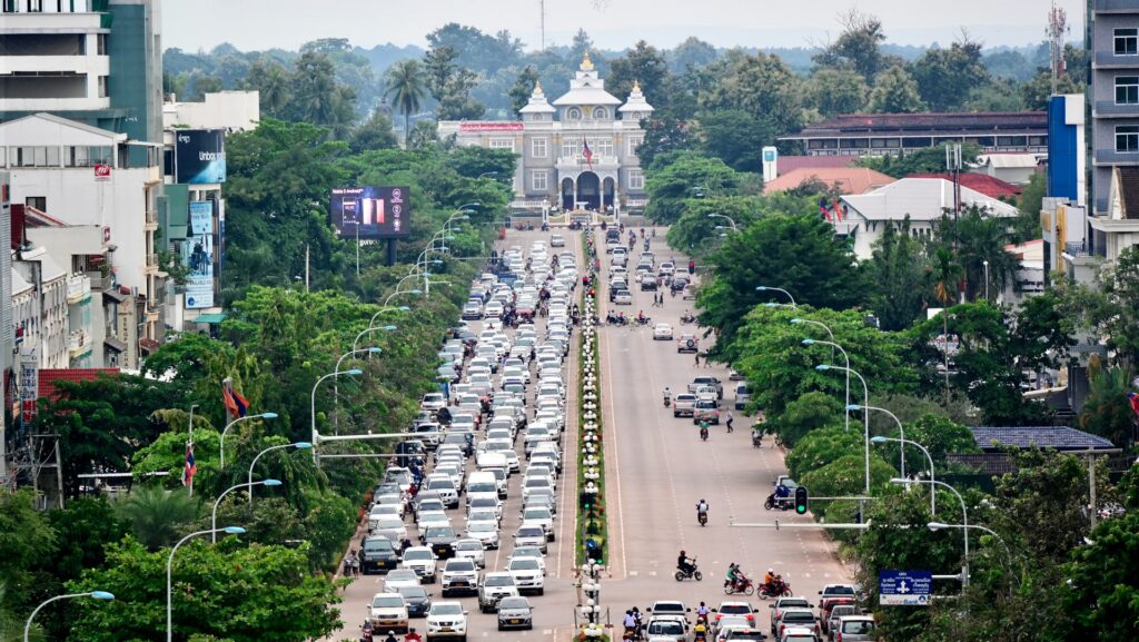 Vientiane, a capital de Laos, uma enorme e extensa avenida nas ruas centrais da cidade, com muitos carros e motos, nas laterais, muita vegetação e árvores, ao fundo, uma construção antiga e as montanhas, para representar o seguro viagem para Laos