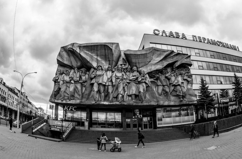 Ruas centrais de Minski com um monumento histórico feito da parte superior de uma loja comercial, com pessoas caminhando lado a lado e olhando para frente, tudo desenhado numa escultura grande de pedra, como se o desenho fosse talhado, para representar o seguro viagem para Belarus