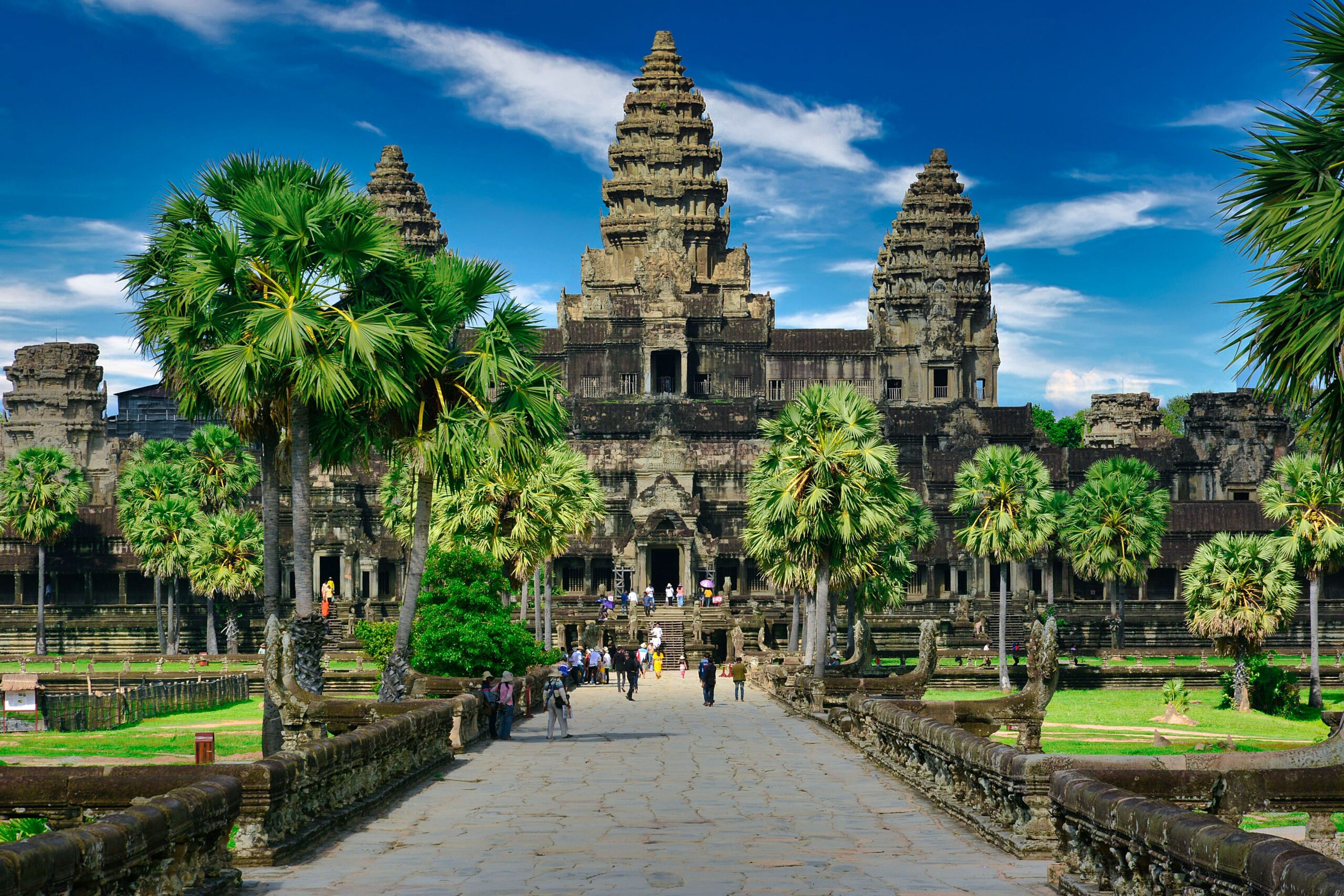 Templo de Angkor Wat no Camboja, com uma passarela de pedra, cercado por palmeiras e vegetação sob um céu azul.