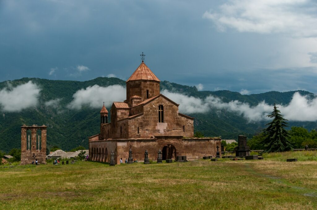 Mosteiro Odzun, uma construção antiga em pedra, bem desgastado ao pé de uma montanha, algumas árvores ao redor, neblina ao fundo, e pessoas visitando o local, para representar o seguro viagem para Armênia