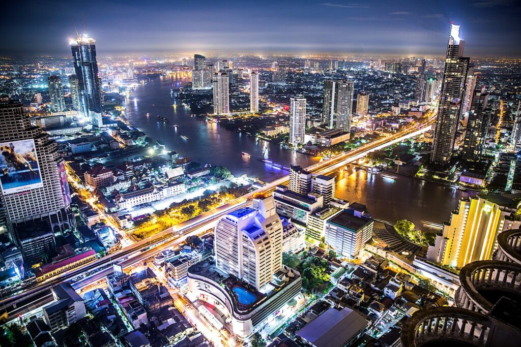 Vista da capital da Tailãndia, Bangkok, com prédios modernos, muitas luzes, uma enorme avenida se estende até uma ponte que corta a cidade, no rio alguns barcos luminosos fazem o percurso, para representar o seguro viagem para Bangkok