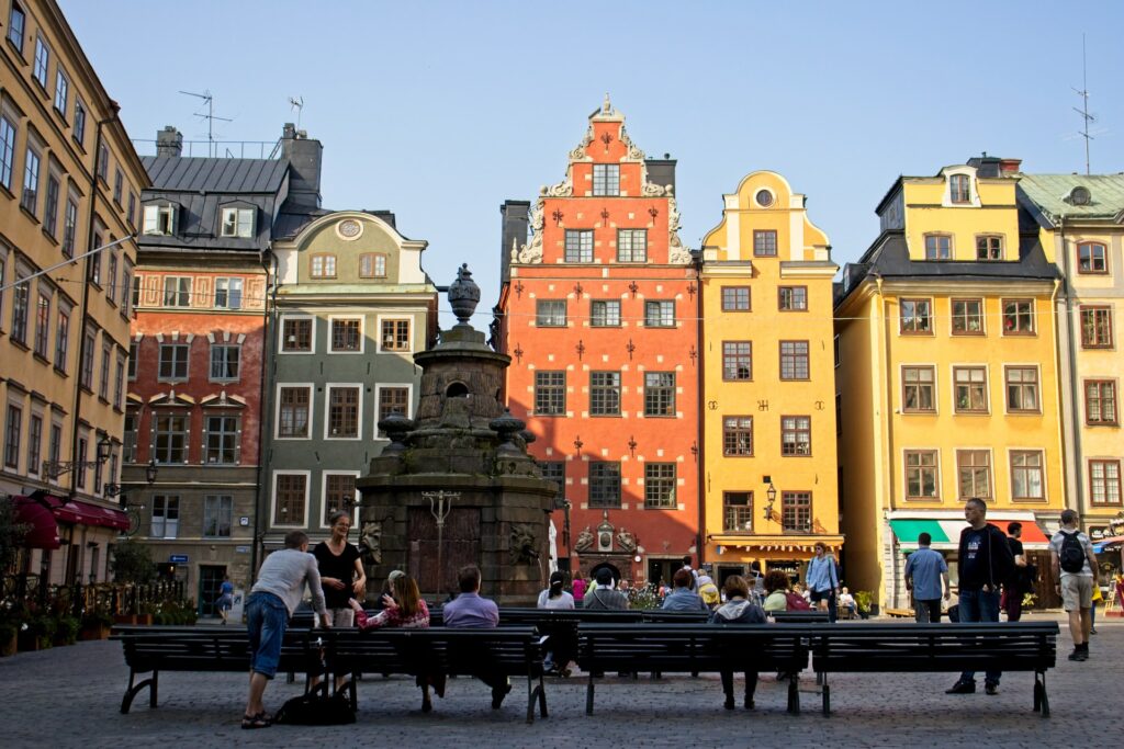 Stortorget, centro histórico, em Estocolmo, com prédios em arquitetura antiga em tons de vermelho e amarelo, uma fonte, e muitos bancos ao redor com pessoas sentadas conversando, para representar o seguro viagem para Estocolmo
