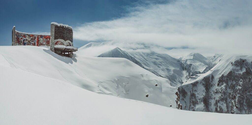 Monumento da Amizade na Cordilheira do Grande Cáucaso na Geórgia, uma construção sem teto, com paredes que formam um círculo, bem na beira de uma montanha completamente coberta por neve com vista para as cordilheiras nevadas, para representar o seguro viagem para Geórgia