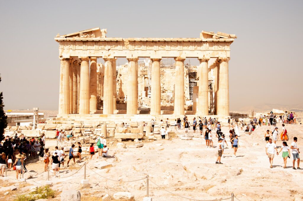 Partenon em Atenas, com oito colunas que sustentam a construção antiga, muitas ruínas ao redor e pessoas caminhando 