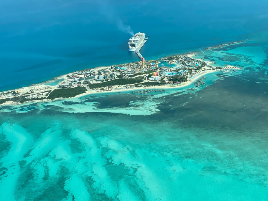 Berry Island nas Bahamas, uma ilha pequena, com um pequeno deck para as embarcações, onde é possível ver um cruzeiro parado, a ilha é repleta de áreas verdes e construções, ao redor, o mar é claro e com corais, para representar o seguro viagem para Bahamas