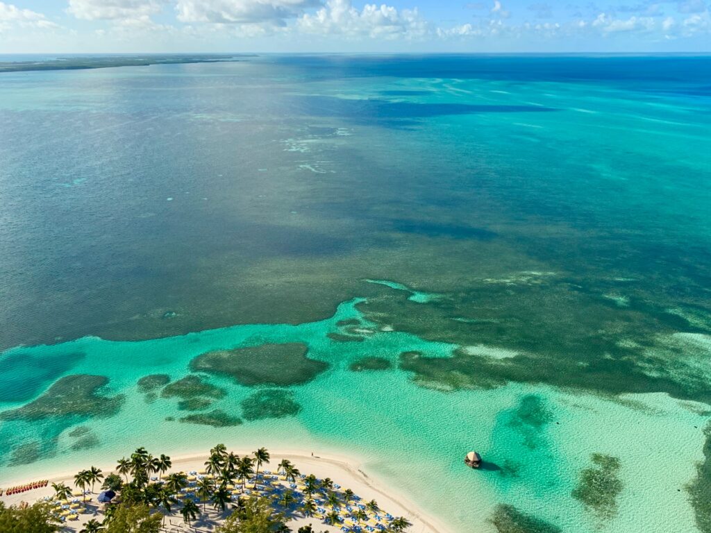 CocoCay, uma das ilhas mais famosas das Bahamas, com foco no mar que começa verde e com muitos corais, e ai escurecendo aos poucos, a faixa de areia é pequena e muito clara, é possível ver guarda-sóis e muitos coqueiros, para representar o seguro viagem para Bahamas